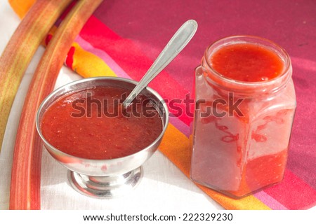 Rhubarb and strawberry classic red jam in a jar and small metallic dish on red background. Rhubarb stalk on the background.