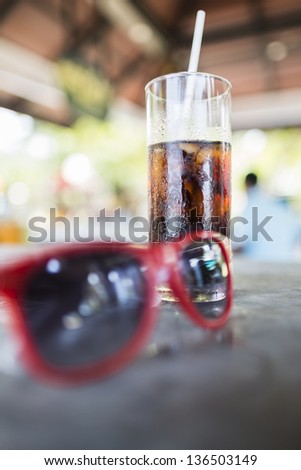 Trendy red sunglasses standing with a glass of iced soda or soft drink in a tall glass on a counter top on a summer day, shallow depth of field