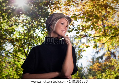 Low angle upper body portrait of a smiling trendy young woman in a cloth cap amongst autumn trees with sun flare