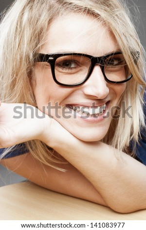 Close-up portrait of a smiling playful blond woman wearing glasses and leaning over her arms and over a desk