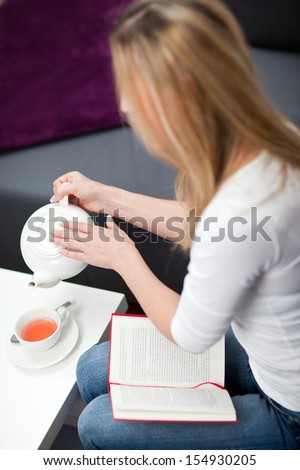 High angle view of a blond woman sitting reading a book stopping and pouring herself a cup of tea from a teapot