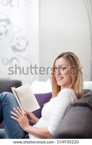 Attractive young woman relaxing in a chair at home reading a book and turning to look at the camera with a beautiful smile