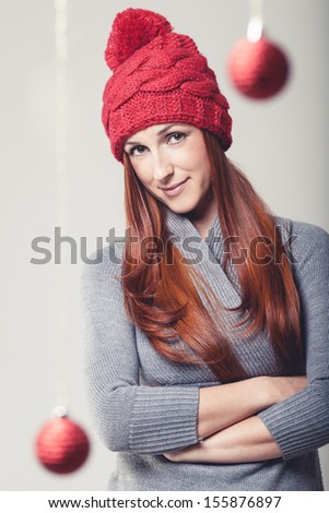 Confident young woman wearing a festive red woolly cap standing with folded arms looking at the camera with a smile with hanging Christmas baubles in the foreground