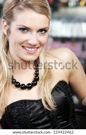 Glamorous beautiful blond woman in an elegant off the shoulder black evening dress and jewelery sitting in a nightclub smiling at the camera, closeup head and shoulders portrait