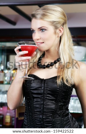 Elegant beautiful young blond woman in a stylish black cocktail dress drinking martini cocktail in a nightclub or bar