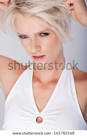 Vindictive angry young blond woman squinting at the camera with slit eyes and a baleful expression as she tears at her hair with her hands, closeup on grey