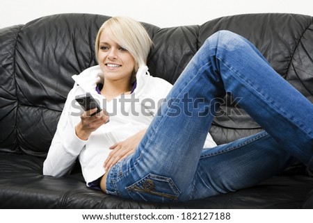 Beautiful woman smiling as she watches television while relaxing on a comfortable black leather sofa in her jeans