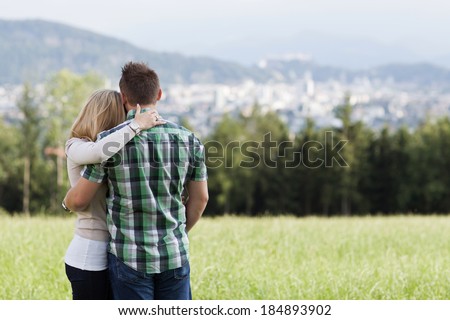 Happy romantic couple standing arm in arm with their backs to the camera in a field looking out over the countryside to a distant town