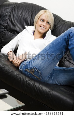 Slender pretty blond woman in jeans relaxing at home reclining on a black leather sofa with a happy smile