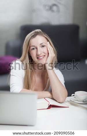 Attractive woman working at home from a home office or studying sitting at a table with a hardcover book and a laptop with her chin on her hand smiling at the camera