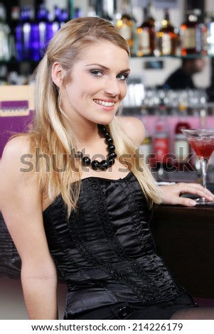 Beautiful stylish blond woman in a nightclub or bar wearing an elegant black cocktail dress and matching necklace leaning against the counter as she enjoys a formal night out