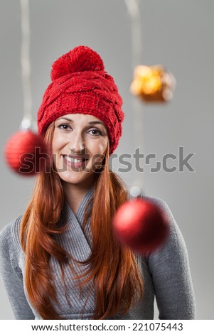 Smiling Pretty Woman in Gray Suit and Red Bonnet Looking at Camera with Christmas Decors Hanging in Front. Isolated on Gray Background.