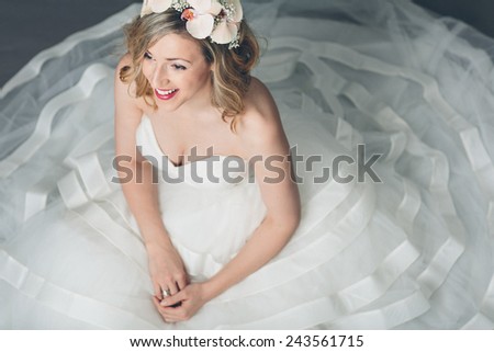 High angle view of an attractive blond elegant young bride in a full skirted white gown sitting on the floor with the fabric spread out round her looking ahead with a happy smile