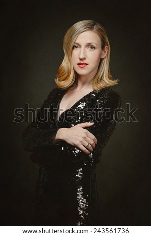 Attractive slender blond woman in a chic black cocktail dress standing sideways with folded arms looking at the camera with a serious expression, over dark grey