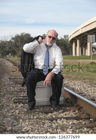 Jobless senior businessman sits on suitcase on railroad train tracks pondering his uncertain future.