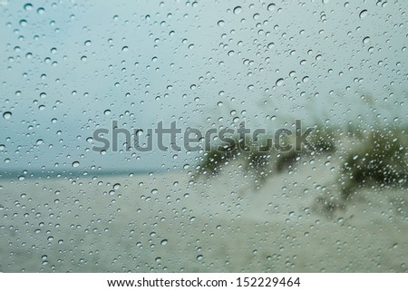 Looking through car windshield with raindrops and rain running down the glass. Beyond is a blurry view of the beach