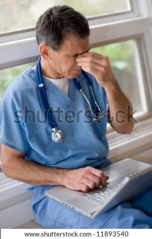 Mature doctor in blue scrubs sitting on floor with laptop, rubbing his tired eyes, looking overworked and tired
