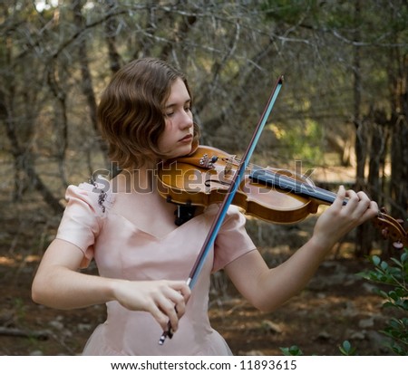 Pretty young woman playing the violin in a forest wearing a pink vintage evening gown. Short depth of field.