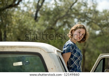 Pretty young woman or teenager standing on running board of vintage  truck looking at viewer, blurred foliage in background