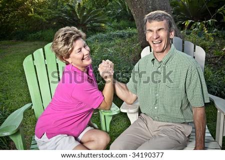 Happy mature couple arm wrestling outdoors. The woman is winning, the man is laughing.