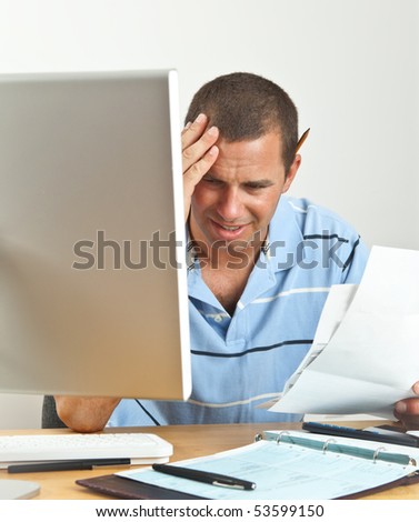 Worried young man holds his head in pain as he wonders how he will pay the bills. Seated at desk with computer, checkbook, stacks of bills and calculator.