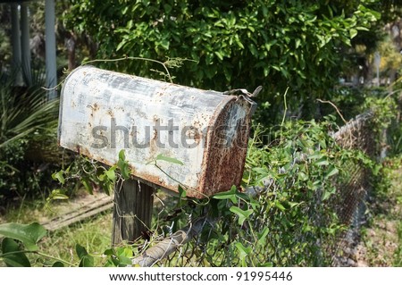 Rusty mailbox next to a chain link fence. Vines and overgrown yard in background at an abandoned house.