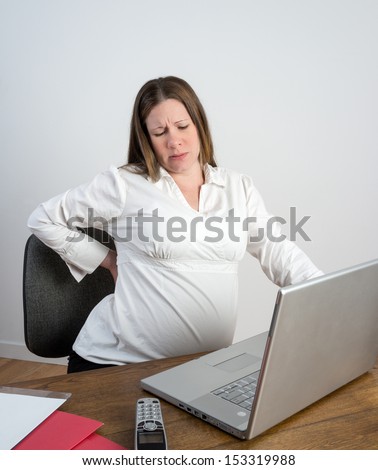 Pregnant young woman rubbing her aching back and stretching at her desk.