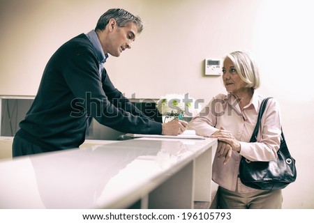 Senior female patient at hospital reception desk with man in 40s collecting personal data.