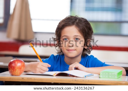 Kid at school writing on his book with an apple on the desk.