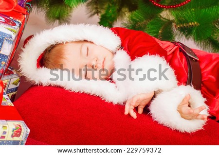 baby in Santa costume sleeping at the Christmas tree with gifts