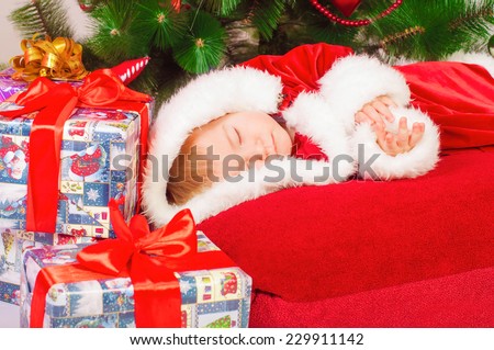 Baby in Santa costume sleeping at the Christmas tree with gifts