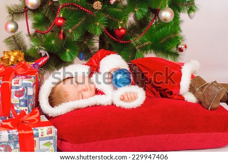Baby in Santa costume sleeping at the Christmas tree with Christmas toy in hands