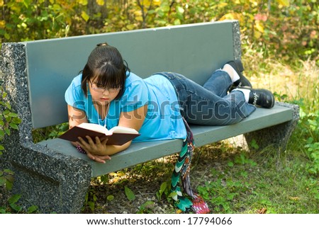 A nine year old girl reading a book outside on a bench