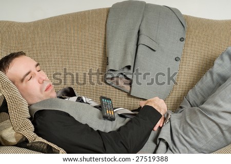 A young businessman sleeping on the sofa with a television remote on his belly