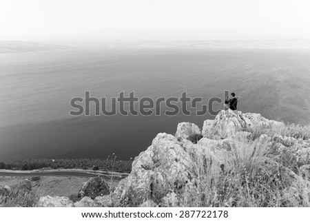 Man photographer sitting taking picture  resting stone cliff above lake sea, Arbel mountain, Kineret Galilee Tiberias lake, Israel. Black and white.