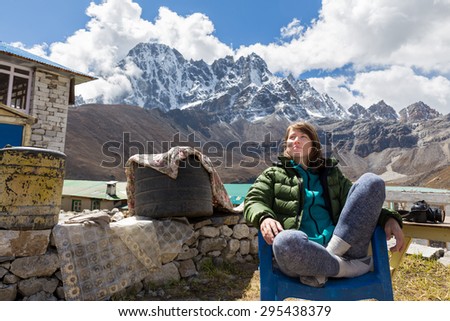 Young smiling happy cheerful woman tourist sitting chair resting . Gokyo village lodges, snow mountains peaks ridge range lake view. Everest Base Camp route trail trekking traveling, Nepal tourism.