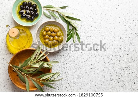 Green and black olives with olive oil in a glass bottle, olive tree sprigs and cut fresh ciabatta bread on wooden cutting board. White rustic background, healthy mediterranean food, space for text