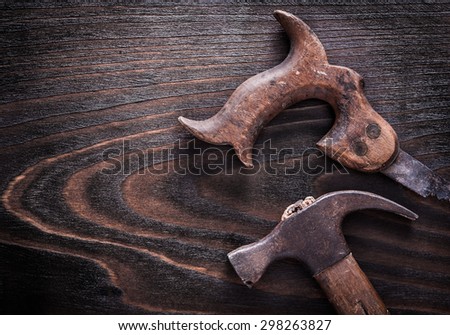 Rusted metal handsaw with claw hammer on vintage dark wooden board close up view construction concept.