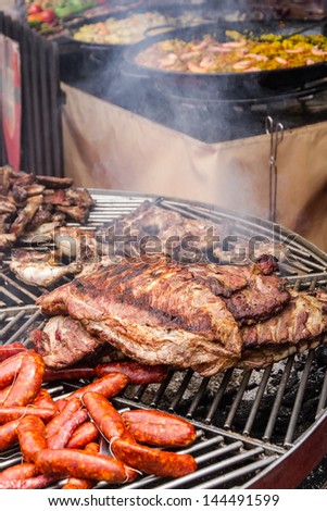 grilled meat and smoke lit outdoor fair. Pork, chicken and beef, sausages. In the background paellas and fruits