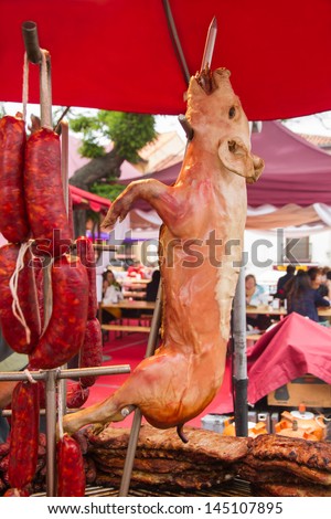 Roast suckling pig ready for consumption by beef rib roast or chops and pork sausages hanging. People in food stall, outdoors, in medieval fair