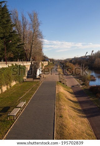 Bike Path and Pedestrian Walkway in city with greenery, shrubs and trees along the river. People walking and sitting in the sun in winter afternoon
