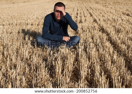 Young man sitting on the floor in mown cereal field, covering with hand the strong sun