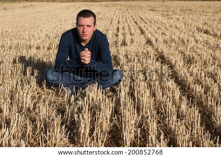 Young man sitting on the floor in mown cereal field with the fingers intertwined hands and looking at camera