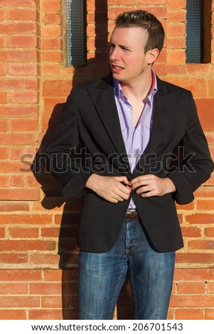 Young man buttoning his jacket in the sun in front of a brick wall