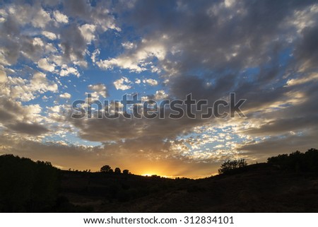 Sunset with the sun shining hidden among hills, trees and the sky full of clouds