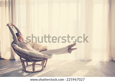 Young woman at home sitting on modern chair in front of window relaxing in her living room reading book, instagram toning 商業照片 © 