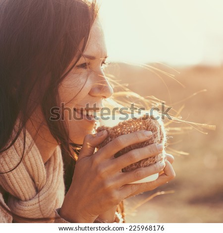 Woman wearing warm knit clothes drinking cup of hot tea or coffee outdoors in sunlight, instagram toned, square composition