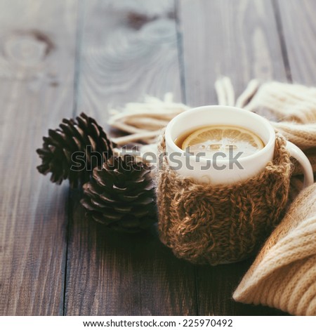 Cup of hot tea with lemon dressed in knitted warm winter scarf on brown wooden tabletop, square photo, soft focus