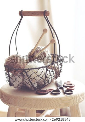 Vintage knitting needles, scissors and yarn inside old wire basket on wooden stool, still life photo with soft focus