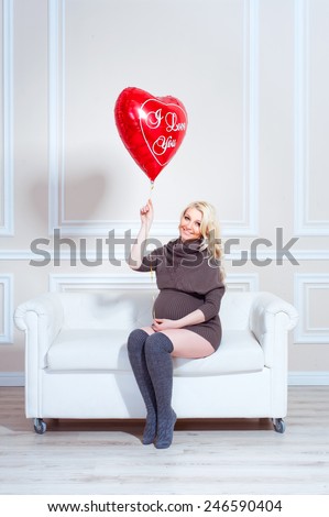 young beautiful pregnant woman sitting on a sofa and holding balloon in the form of heart in hand. Valentine\'s Day.